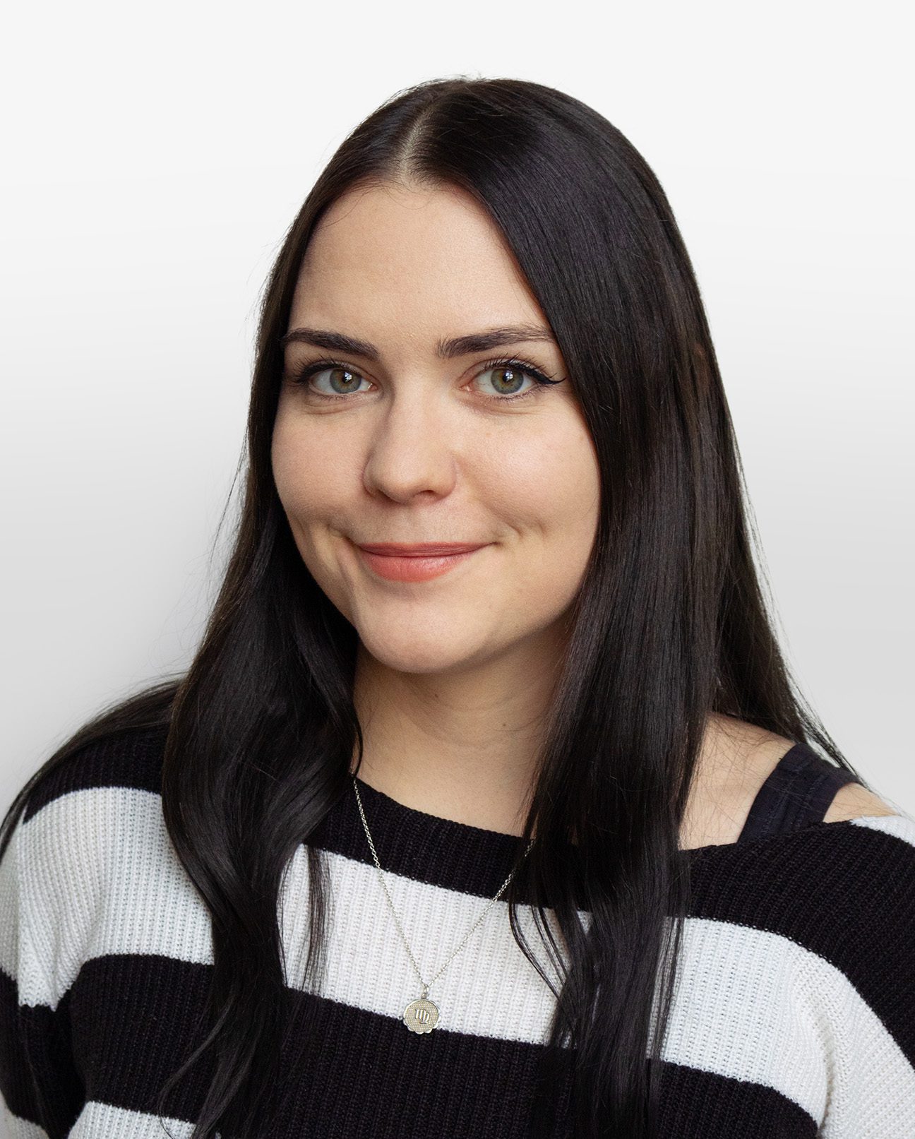 A woman with long straight black hair, light skin, and blue eyes smiles slightly. She wears a black and white striped sweater and a silver necklace, posed against a plain white background—perfect for Vu, le plus grand inventaire de montures à Montréal.