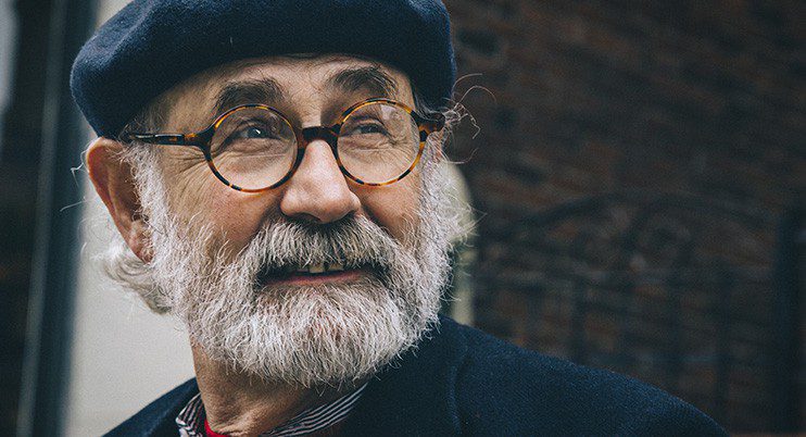 An older man with a white beard and round glasses from Vu, le plus grand inventaire de montures à Montréal, wears a dark beret and coat, glancing to the side with a gentle smile against a blurred brick wall.