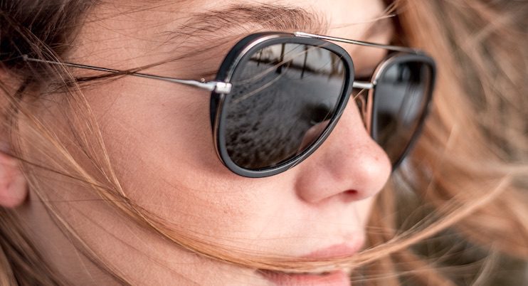 Close-up of a person in black aviator sunglasses from Vu, renowned for having le plus grand inventaire de montures à Montréal. Light brown hair blows across their face, and a vibrant beach scene is reflected in the lenses.