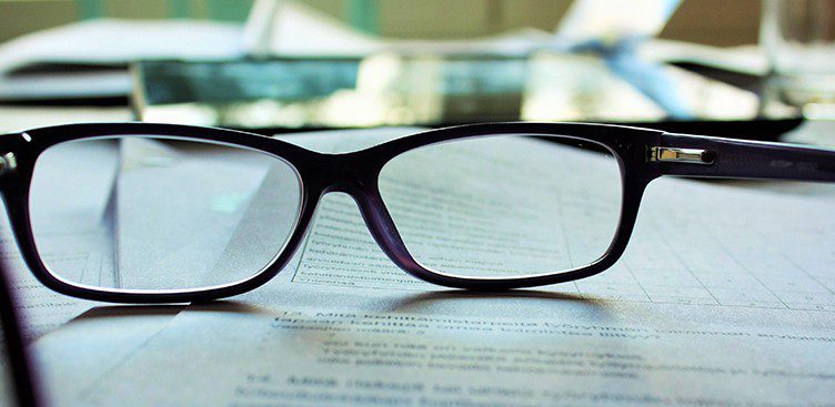 A close-up of eyeglasses from Vu, le plus grand inventaire de montures à Montréal, resting on a document with printed text; the focus is on the lenses and frame, with blurred papers and objects in the background.