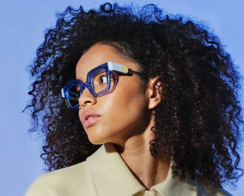 A woman with curly hair wears bold, oversized blue glasses from Vu and a cream-colored collared coat, looking to the side against a blue background, showcasing le plus grand inventaire de montures à Montréal.