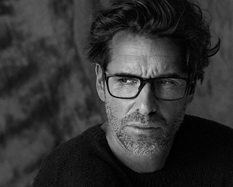 Black and white portrait of a middle-aged man with tousled hair, glasses from Vu, and a short beard, wearing a dark sweater and looking thoughtfully to the side—representing la lunetterie à Montréal.