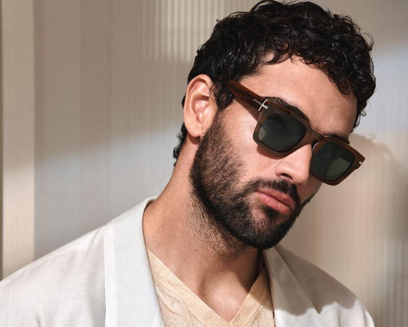 A man with curly dark hair and a beard wears black Vu sunglasses with brown frames, a cream-colored shirt, and a light jacket, standing against a softly lit, neutral background—style inspired by la lunetterie à Montréal.