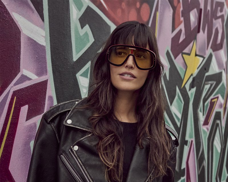 A woman with long dark hair, wearing large yellow Vu sunglasses and a black leather jacket, stands in front of a colorful graffiti wall.