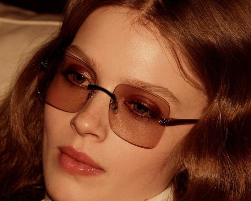 A person with long brown hair wearing large, square, tinted glasses from Vu looks downward with a calm expression. The warm lighting highlights smooth skin and natural makeup, reflecting the style found at la lunetterie à Montréal.
