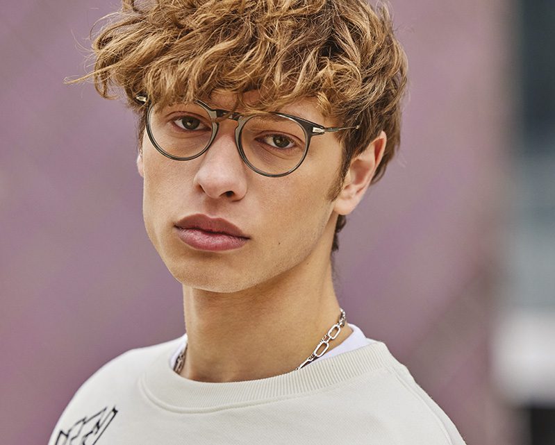 A young person with wavy, light brown hair and round glasses from Vu lunetterie à Montréal looks into the camera. They wear a white sweatshirt and a silver chain necklace, with a blurred background behind them.
