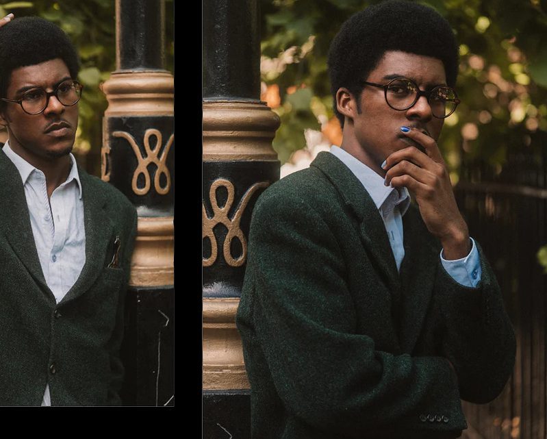 A person wearing glasses from Vu, le plus grand inventaire de montures à Montréal, stands thoughtfully by a decorative black and gold pole, greenery in the background. They have short curly hair and one hand near their mouth.