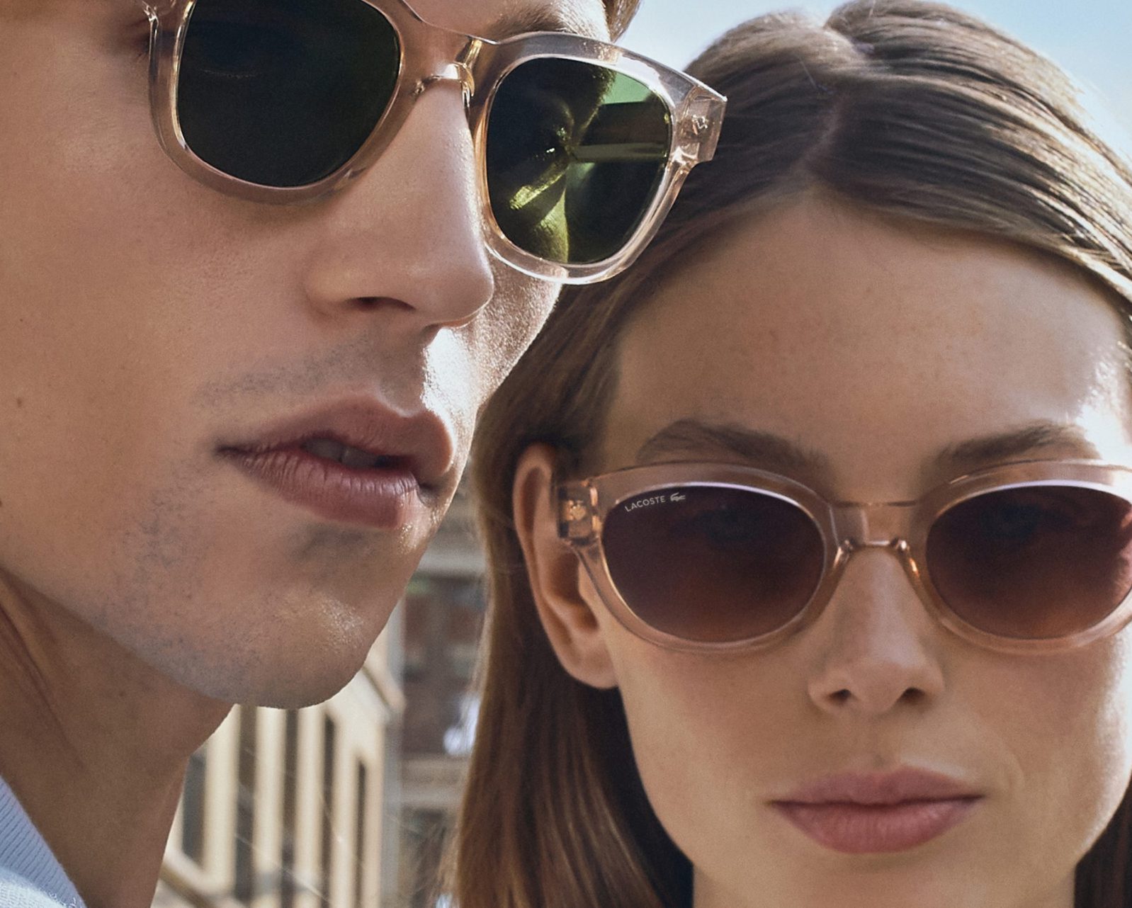Close-up of a man and woman wearing Vu sunglasses outdoors, with buildings blurred in the background. Both have neutral expressions as sunlight highlights their faces, showcasing the chic style from le plus grand inventaire de montures à Montréal.