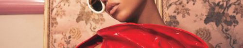 A person wearing oversized white sunglasses from Vu lunetterie à Montréal and a shiny, sculptural red outfit with a yellow and white beaded detail stands in front of a vintage floral wallpaper.