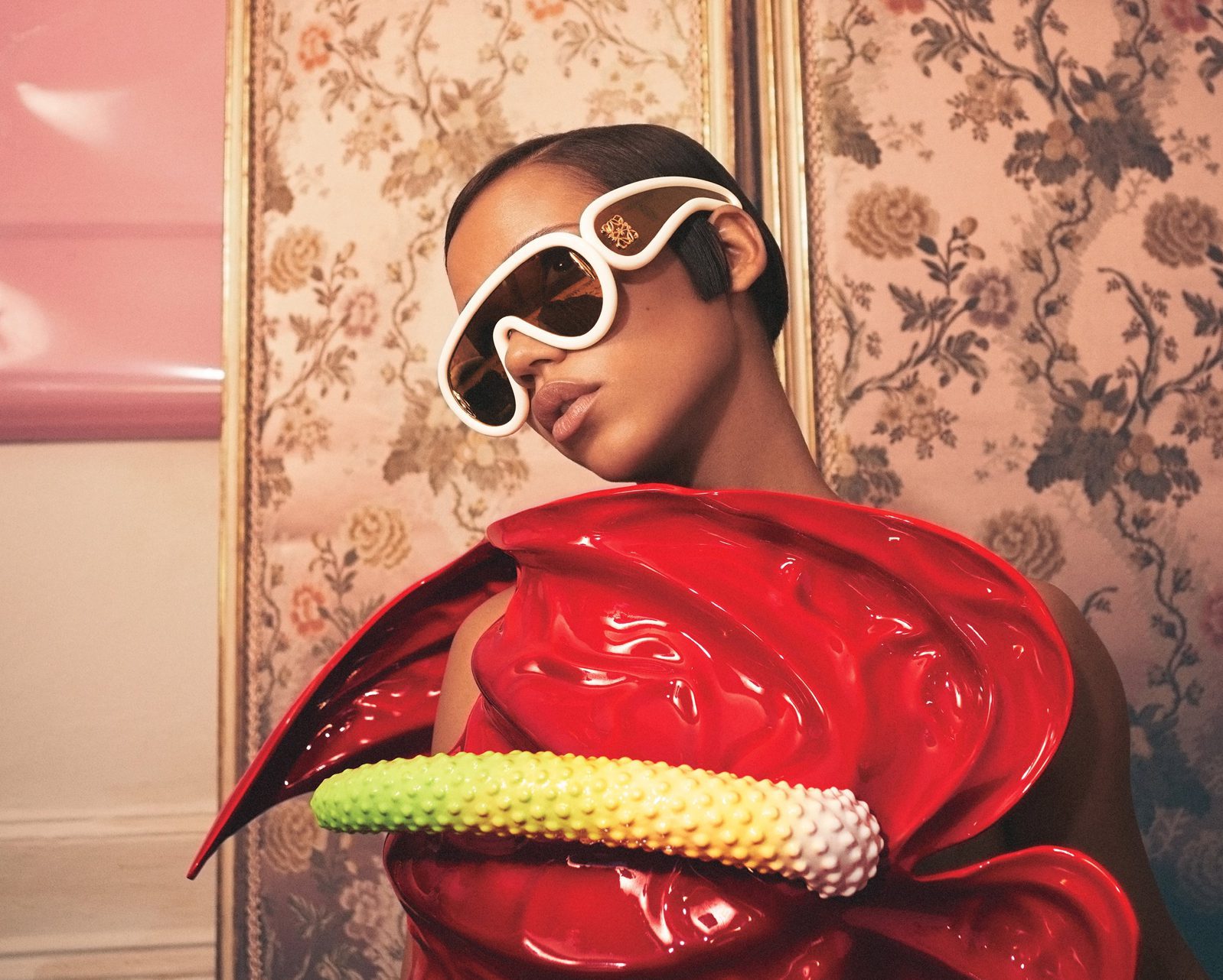 A person wearing oversized white sunglasses from Vu lunetterie à Montréal and a shiny, sculptural red outfit with a yellow and white beaded detail stands in front of a vintage floral wallpaper.