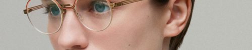 A person with short, dark hair and clear skin wears round, gold-rimmed glasses from Vu, le plus grand inventaire de montures à Montréal, looking slightly to the side with a neutral expression against a plain light background.