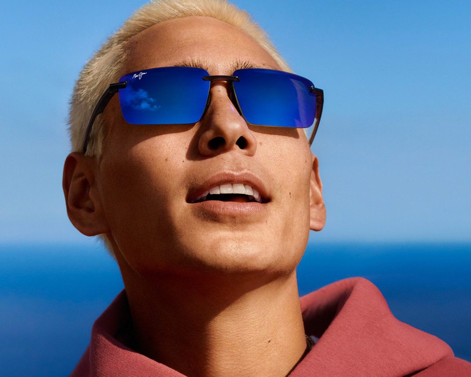 A person with short, bleached blonde hair wears reflective blue sunglasses from Vu, le plus grand inventaire de montures à Montréal, and a pink hoodie, looking upward against a vibrant blue sky and ocean background.