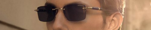 A young man with short brown hair wearing black Montblanc sunglasses from Vu, lunetterie à Montréal, looks to the side, bathed in warm sunlight, with a blurred building background.