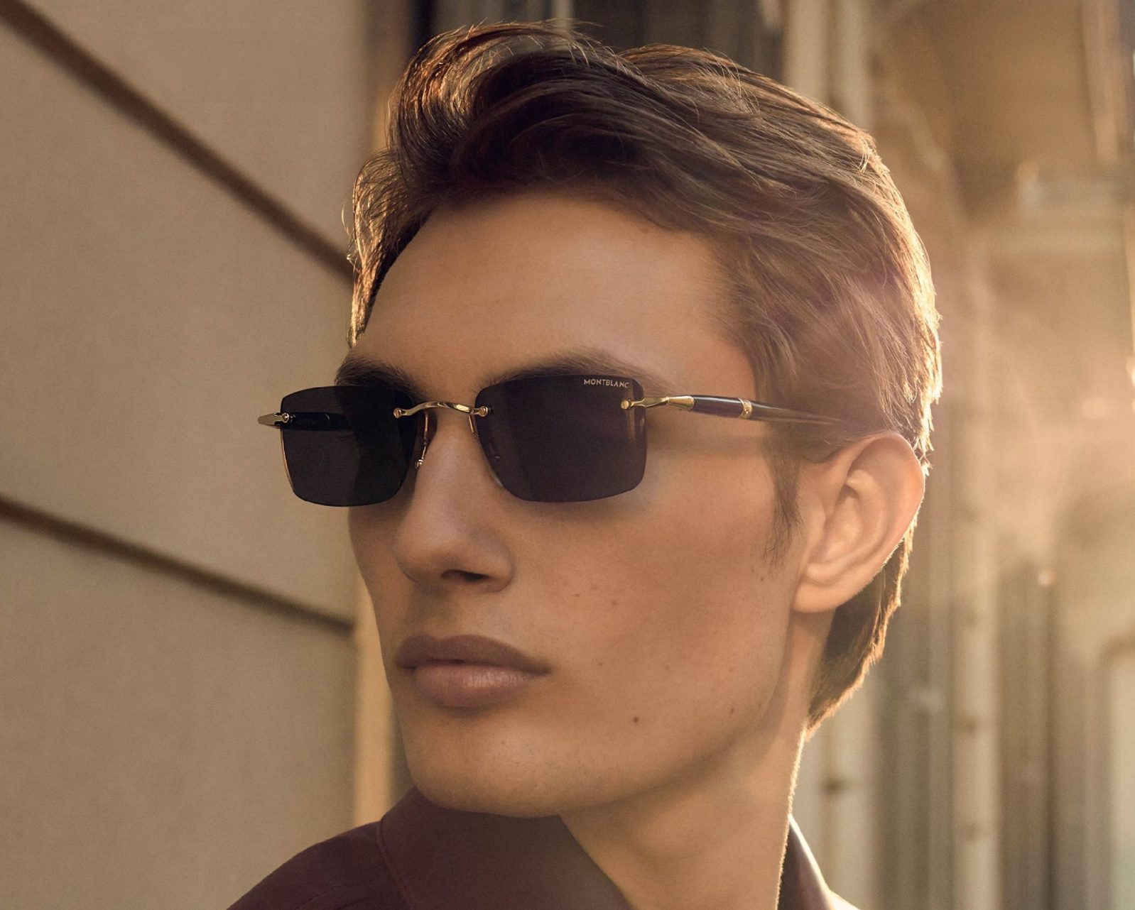 A young man with short brown hair wearing black Montblanc sunglasses from Vu, lunetterie à Montréal, looks to the side, bathed in warm sunlight, with a blurred building background.