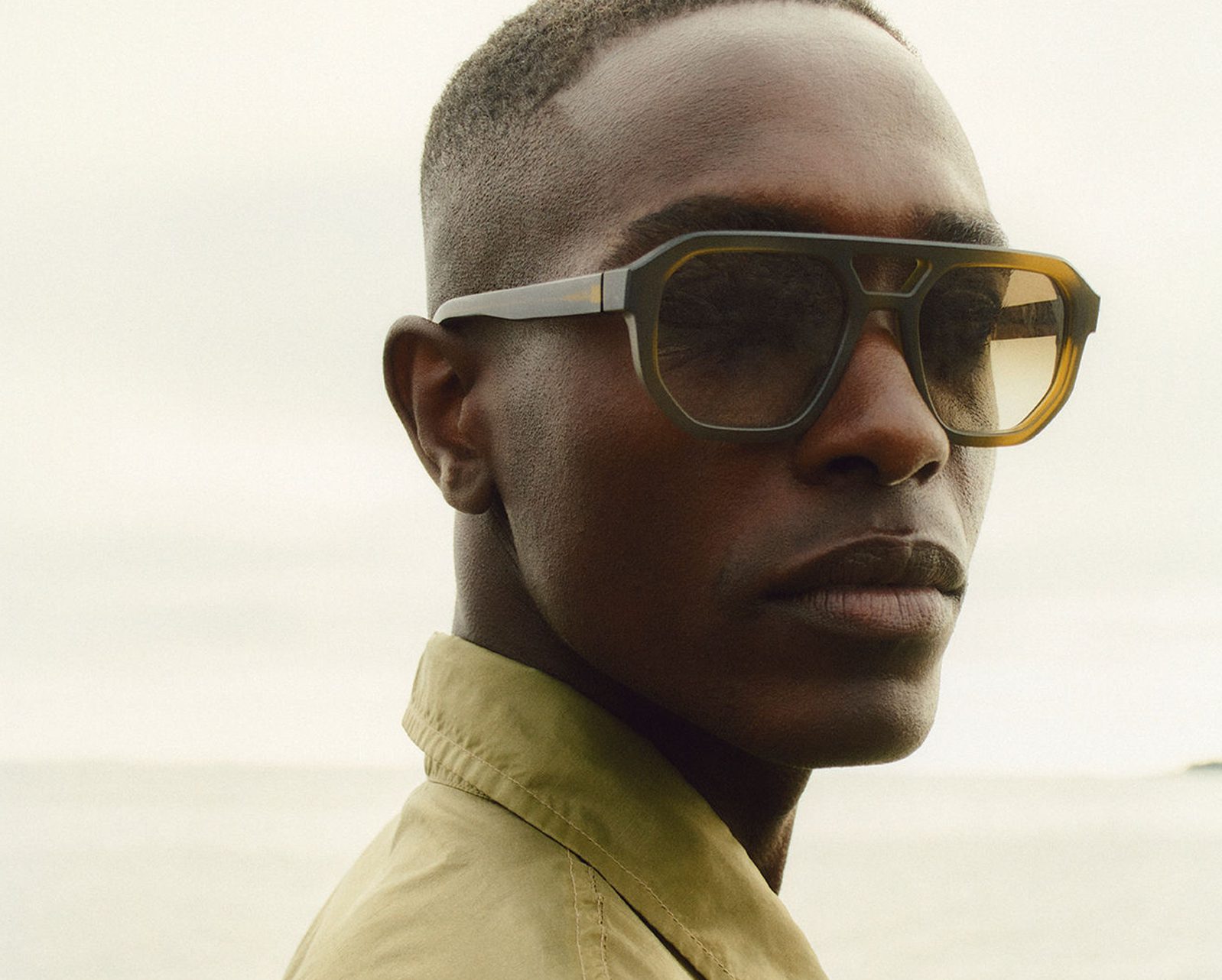 A person wearing large, stylish sunglasses from Vu, le plus grand inventaire de montures à Montréal, and a khaki-colored jacket looks confidently toward the camera with a neutral background, possibly outdoors near water.