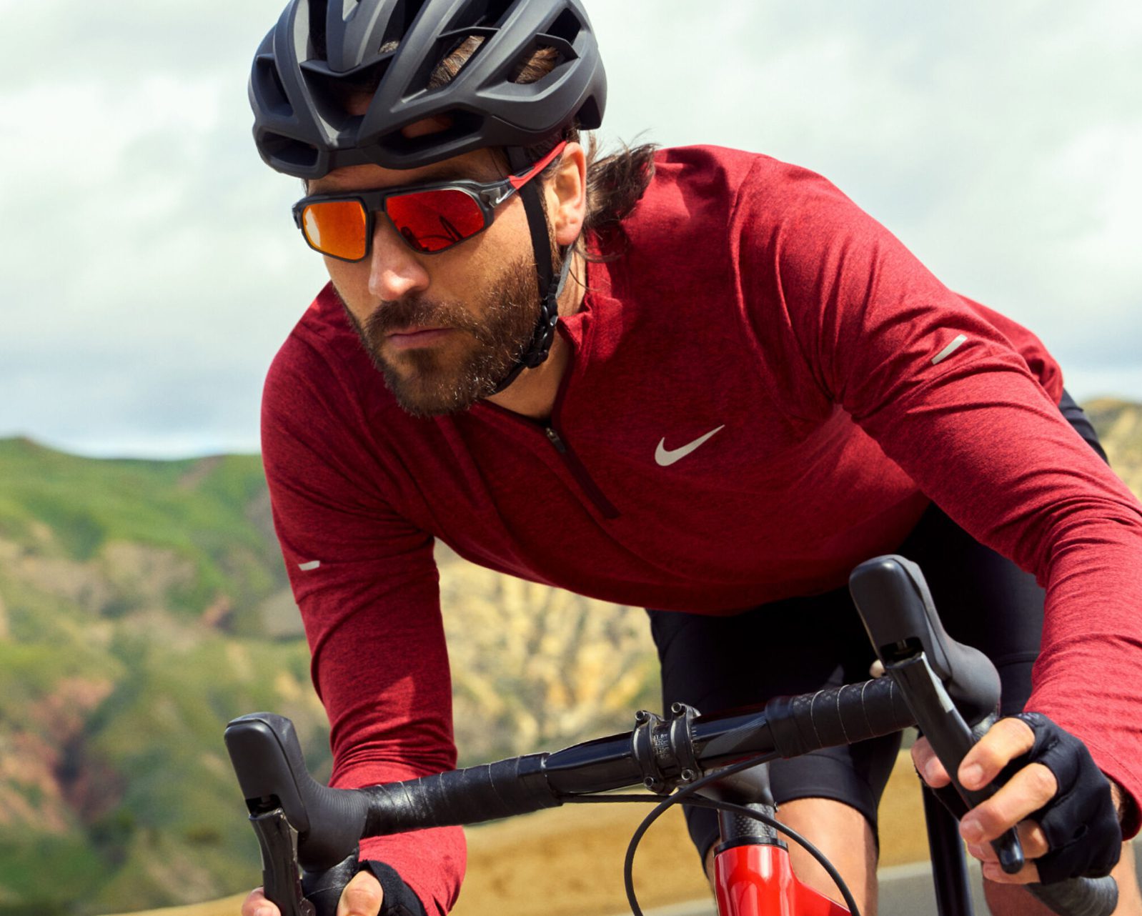 A man wearing a red cycling jersey, black helmet, and reflective Vu sunglasses rides a road bike outdoors with hills and a cloudy sky in the background.