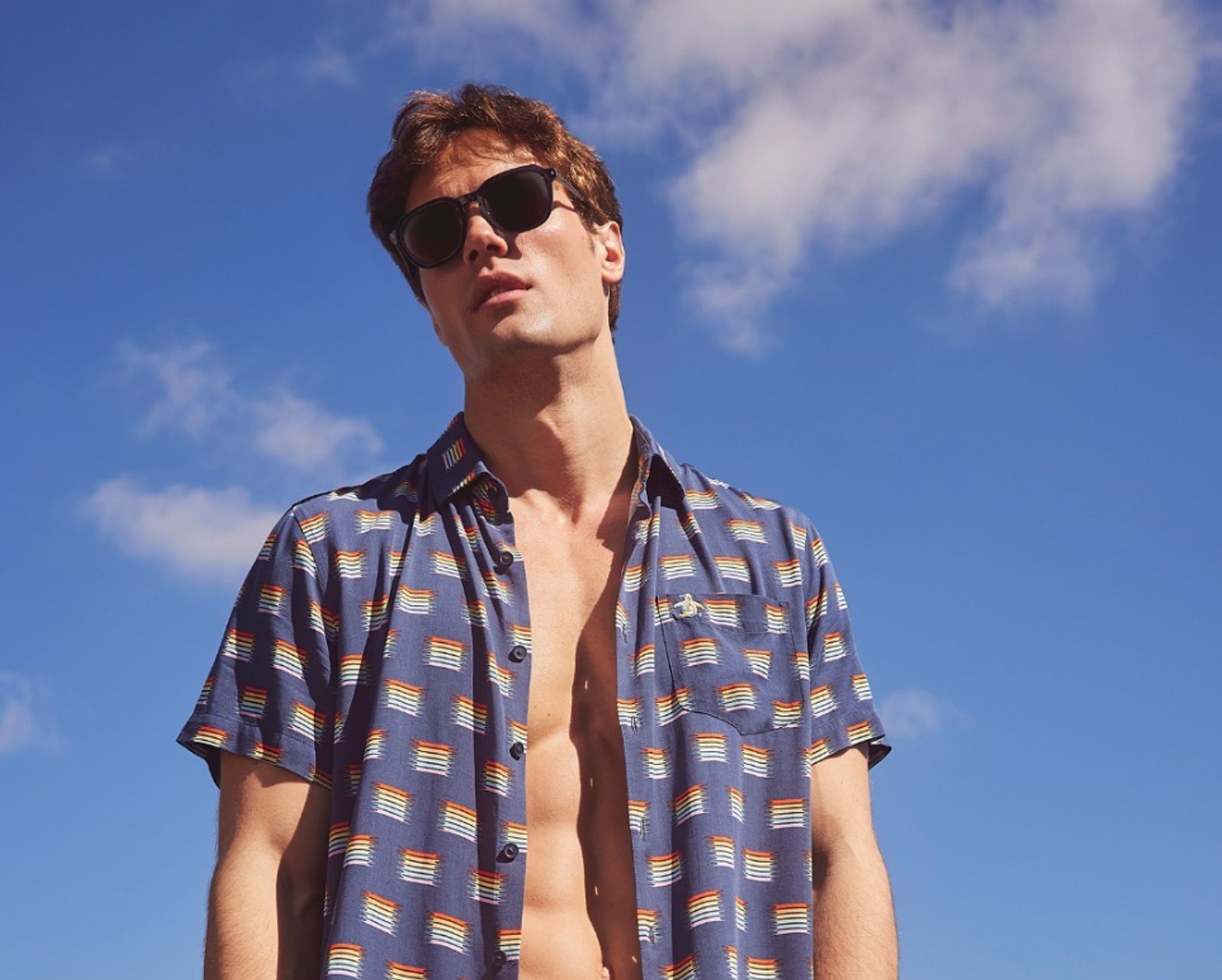 A man wearing sunglasses and an open patterned shirt stands outdoors against a bright blue sky with a few white clouds, showcasing le plus grand inventaire de montures à Montréal.