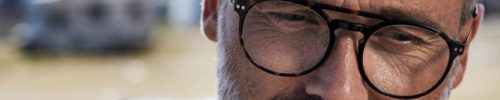 A middle-aged man with gray hair and a beard wears black-rimmed glasses from a lunetterie à Montréal, looking thoughtfully downward. The blurred background hints at sunlight and an outdoor setting.