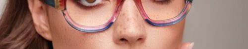 A close-up of a woman with light brown hair wearing large, round, multicolored glasses from Vu, lunetterie à Montréal. She has natural makeup, light brown lipstick, and freckles across her nose, with her hand gently touching her cheek.