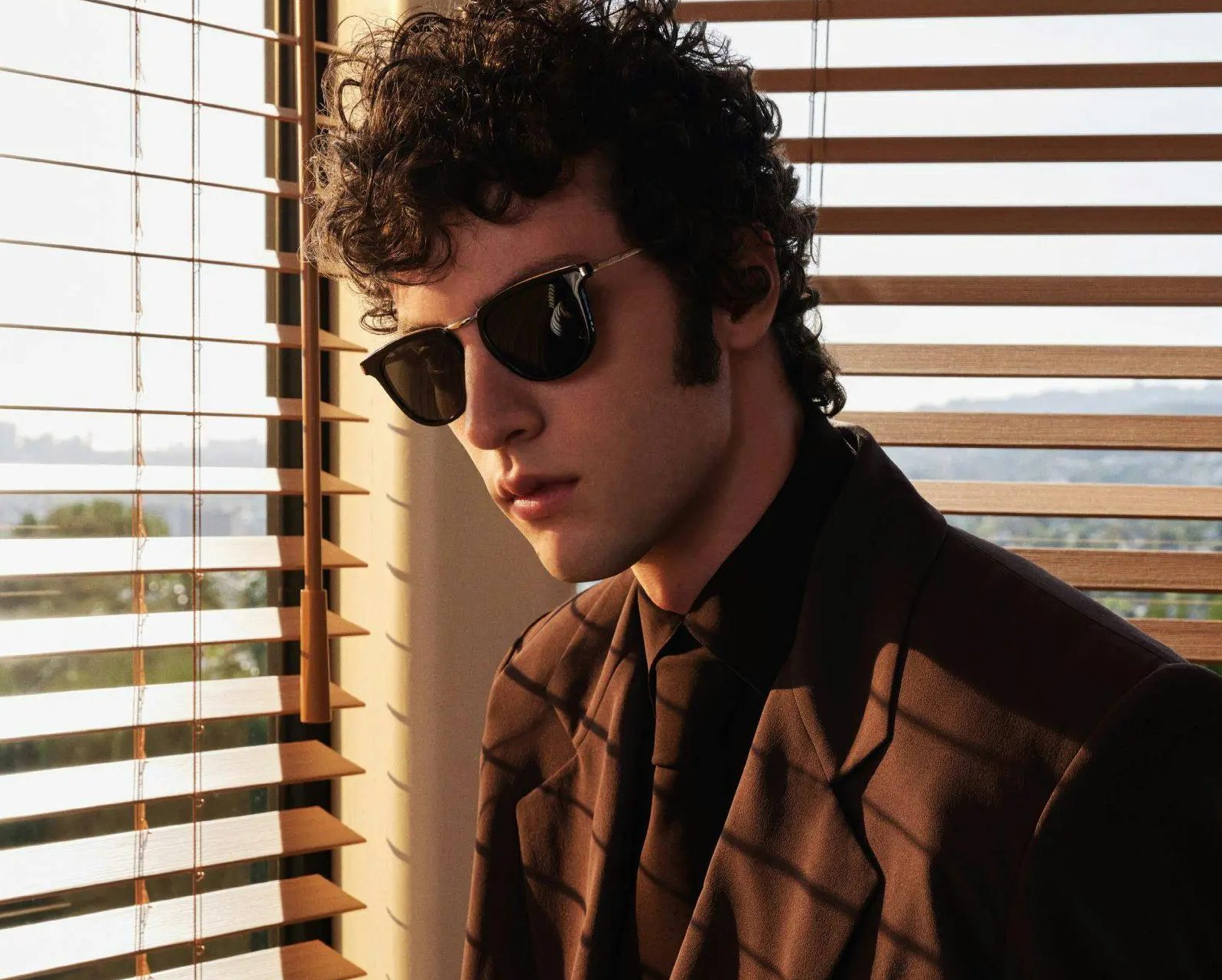 A young man with curly dark hair and sideburns wears black sunglasses from Vu, la lunetterie à Montréal, and a brown suit with a black shirt and tie, standing near window blinds with sunlight streaming through.