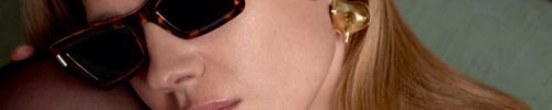 A woman with long blonde hair wearing tortoiseshell sunglasses from Vu, gold heart-shaped earrings, and a patterned top rests her face against her knee, gazing thoughtfully to the side—capturing the style found at la lunetterie à Montréal.