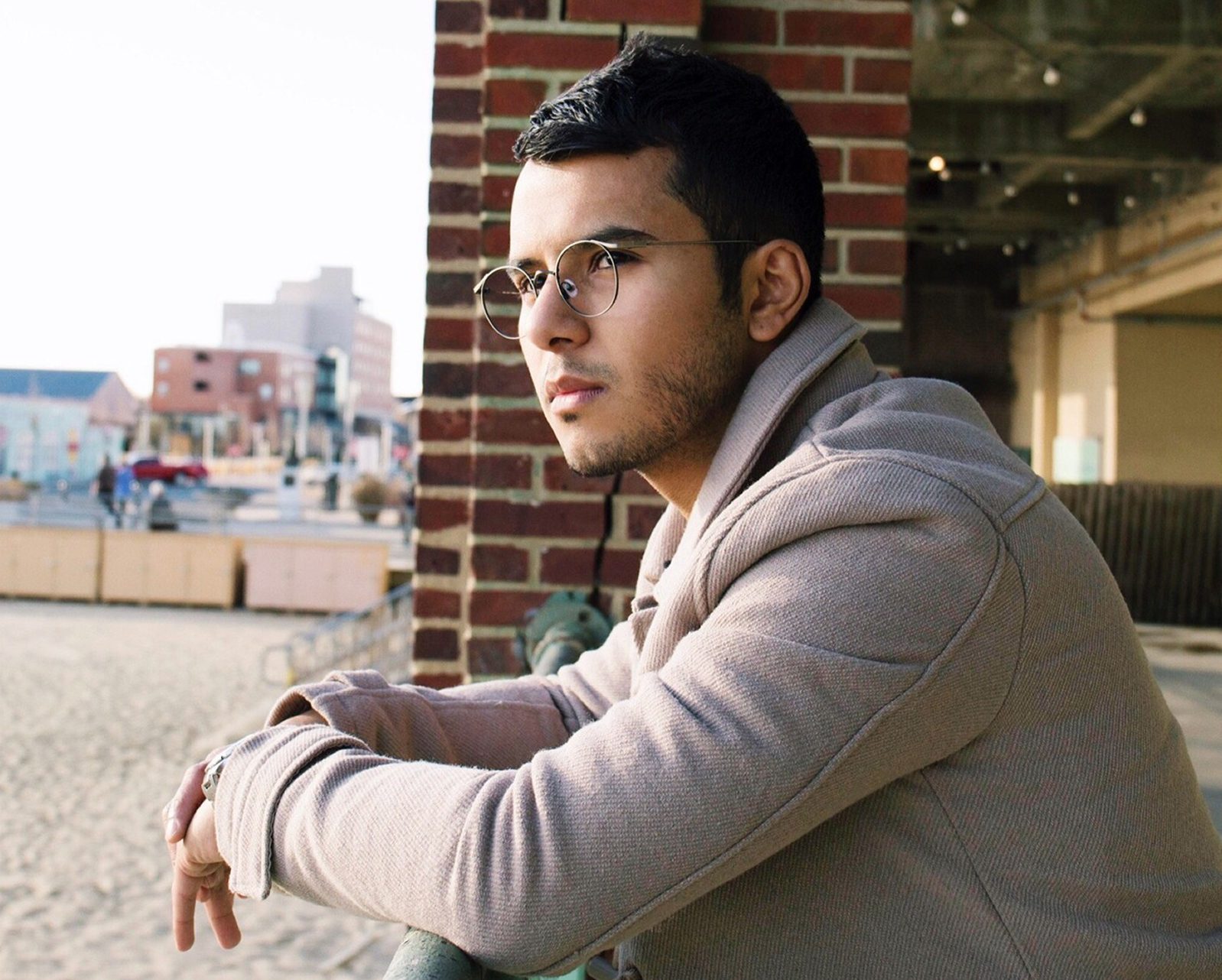 A young man with short dark hair and glasses from une lunetterie à Montréal leans on a railing, gazing thoughtfully into the distance. He wears a light beige coat amid brick buildings, sand, and an urban seaside setting.