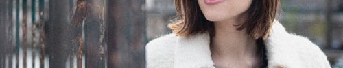 A woman with shoulder-length brown hair and round glasses, wearing a white fuzzy coat over a black top, stands outdoors near a metal fence, looking to the side and smiling softly—capturing the chic style of lunetterie à Montréal.