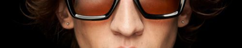 A person with shaggy brown hair wears oversized, thick black sunglasses with amber-tinted lenses—available at Vu, le plus grand inventaire de montures à Montréal—and a gold textured jacket, facing forward against a black background.