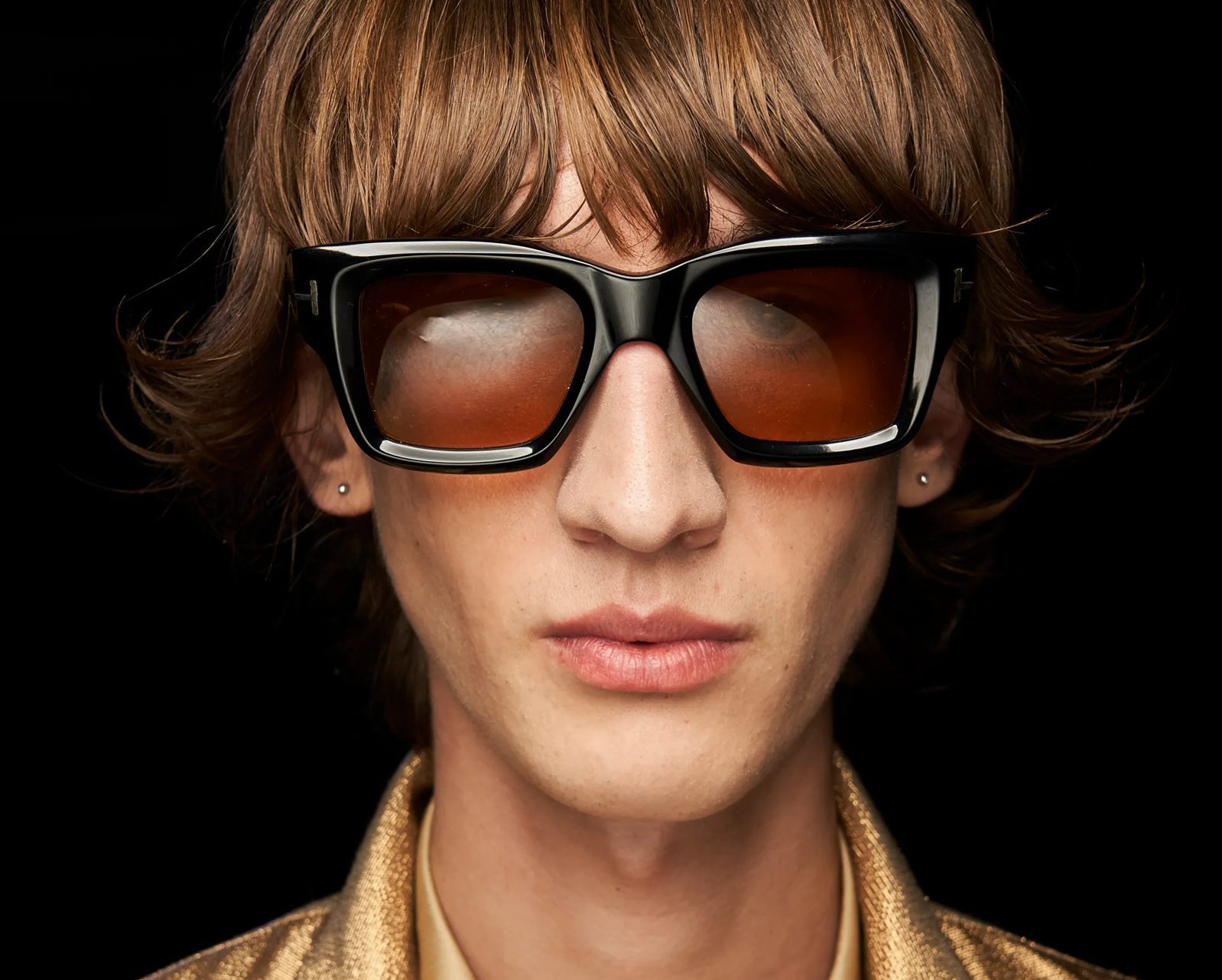 A person with shaggy brown hair wears oversized, thick black sunglasses with amber-tinted lenses—available at Vu, le plus grand inventaire de montures à Montréal—and a gold textured jacket, facing forward against a black background.
