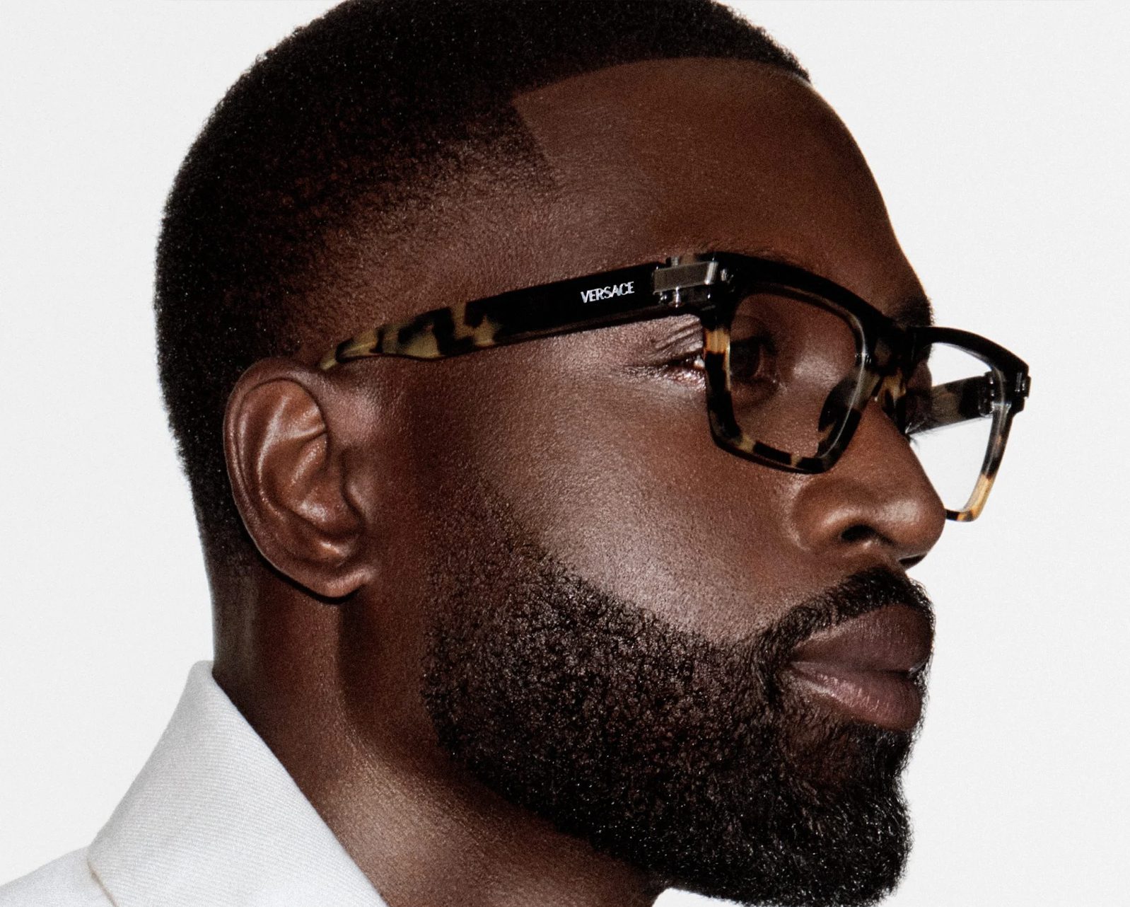A man with a neatly groomed beard and short haircut wears tortoiseshell Versace glasses from Vu, lunetterie à Montréal, and a white shirt, looking to the side against a plain white background.