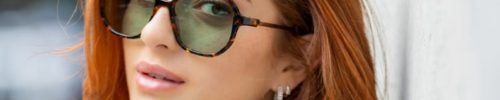 A woman with wavy red hair, wearing dark sunglasses and a black top, leans against a light-colored wall, looking at the camera. Discover le plus grand inventaire de montures à Montréal à Vu, votre lunetterie à Montréal. The background has hints of greenery.
