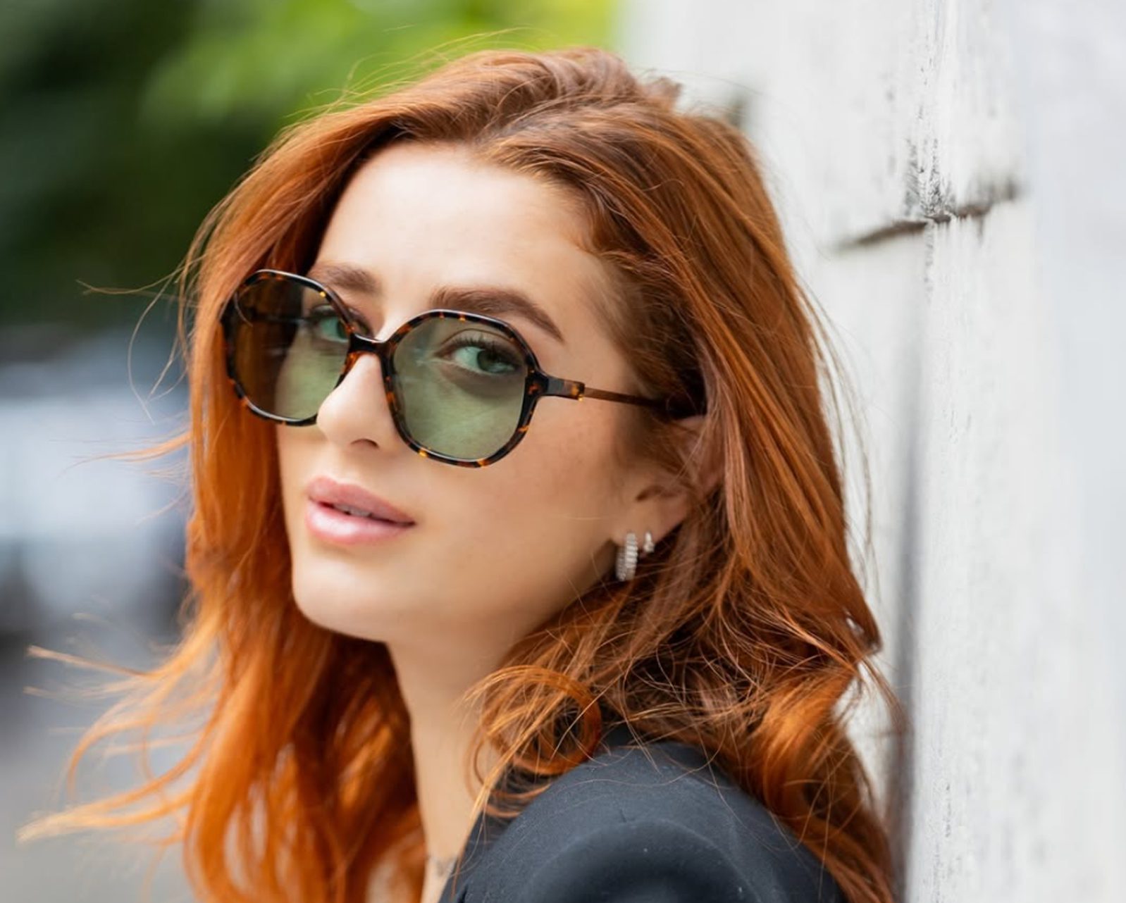 A woman with wavy red hair, wearing dark sunglasses and a black top, leans against a light-colored wall, looking at the camera. Discover le plus grand inventaire de montures à Montréal à Vu, votre lunetterie à Montréal. The background has hints of greenery.