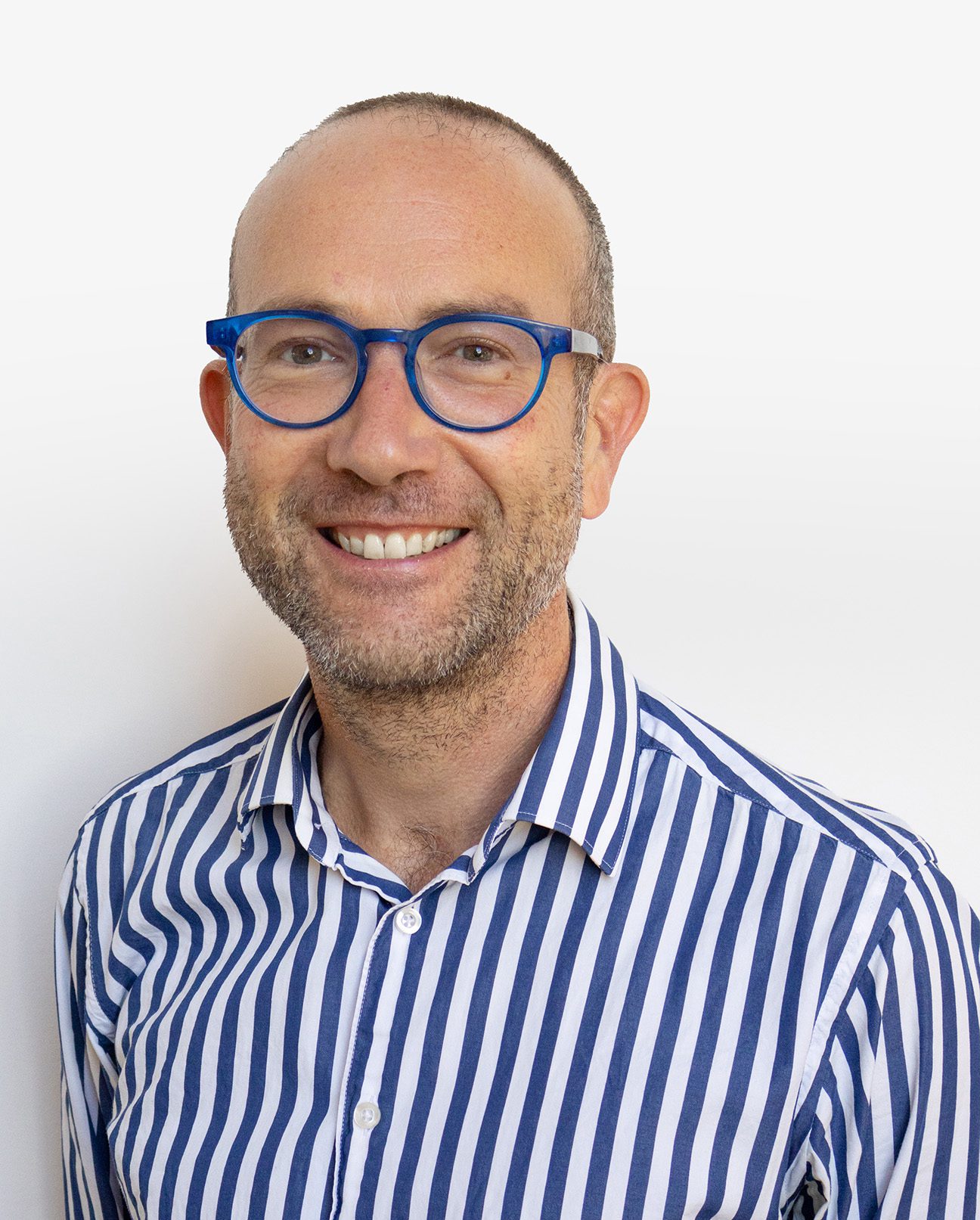 A smiling man with short hair and a beard, wearing blue glasses from Vu, stands in front of a plain white background—showcasing style from le plus grand inventaire de montures à Montréal.