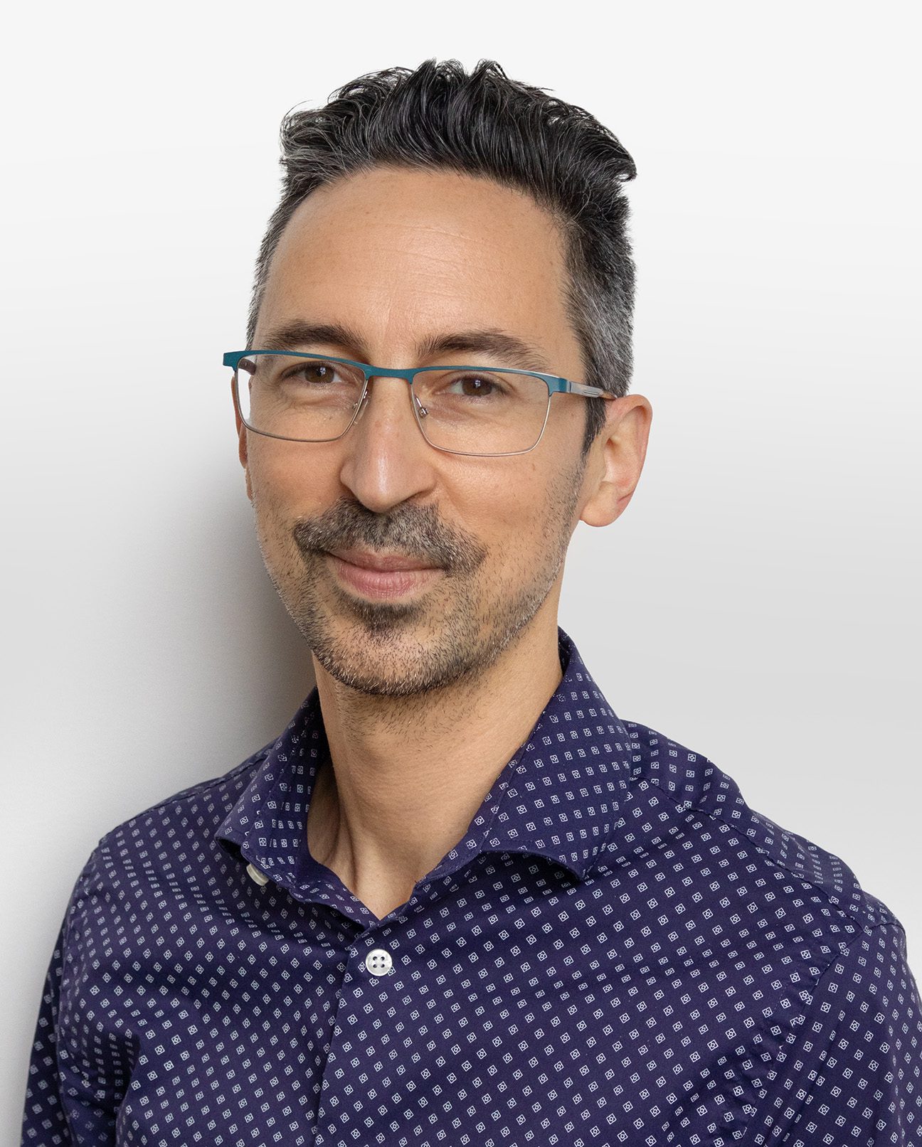 A man with short, dark hair and glasses from Vu, wearing a navy blue button-up shirt with a white geometric pattern, smiles slightly while standing against a plain, light background—showcasing le plus grand inventaire de montures à Montréal.