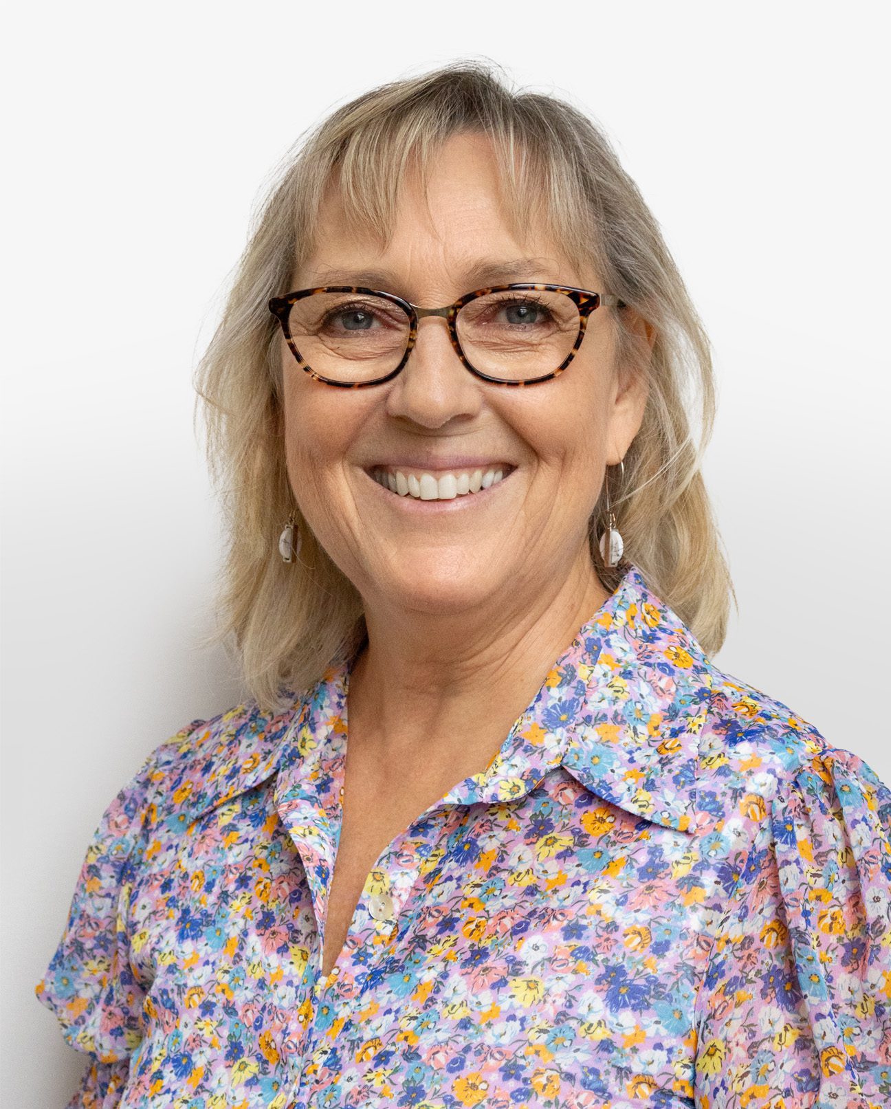 A smiling middle-aged woman with blonde hair, wearing glasses from Le plus grand inventaire de montures à Montréal, pearl earrings, and a colorful floral blouse, stands in front of a plain white background.