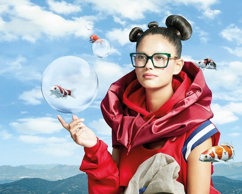 A woman in glasses from Vu stands against a blue sky with clouds, holding a bubble containing a fish. Other bubbles and koi fish float around her, mountains visible behind—a whimsical scene inspired by lunetterie à Montréal.